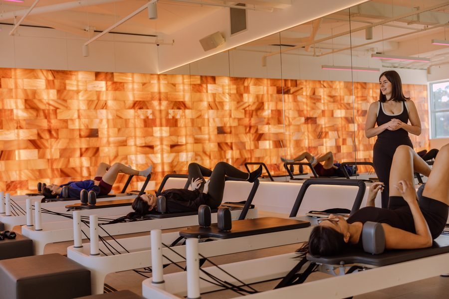Reformer class lying back mid exercise, instructor watching, salt wall along the room
