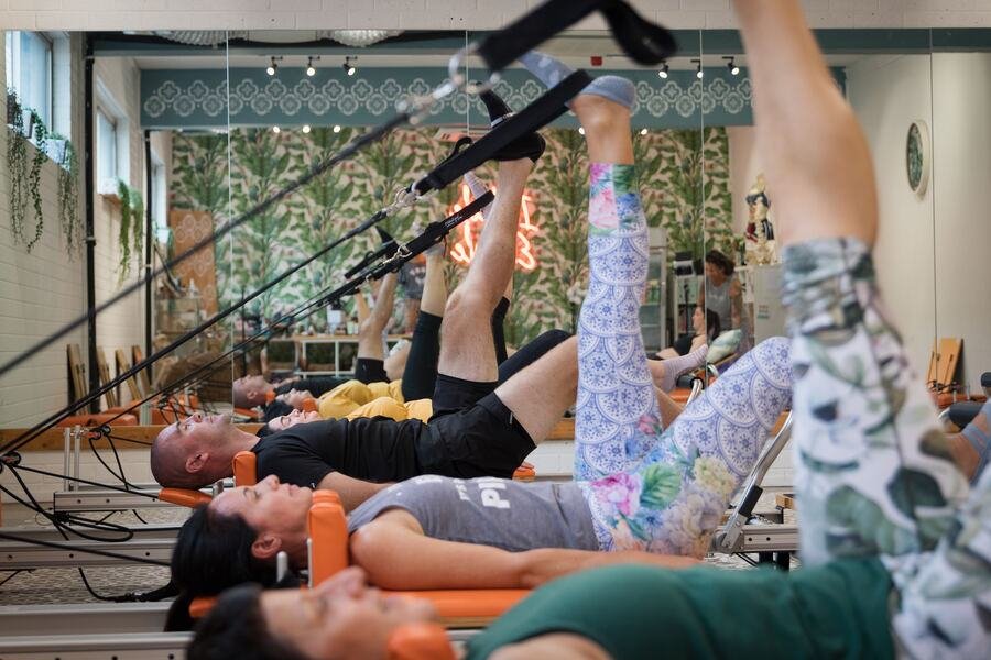 Members lying back on orange reformers with arms extended overhead