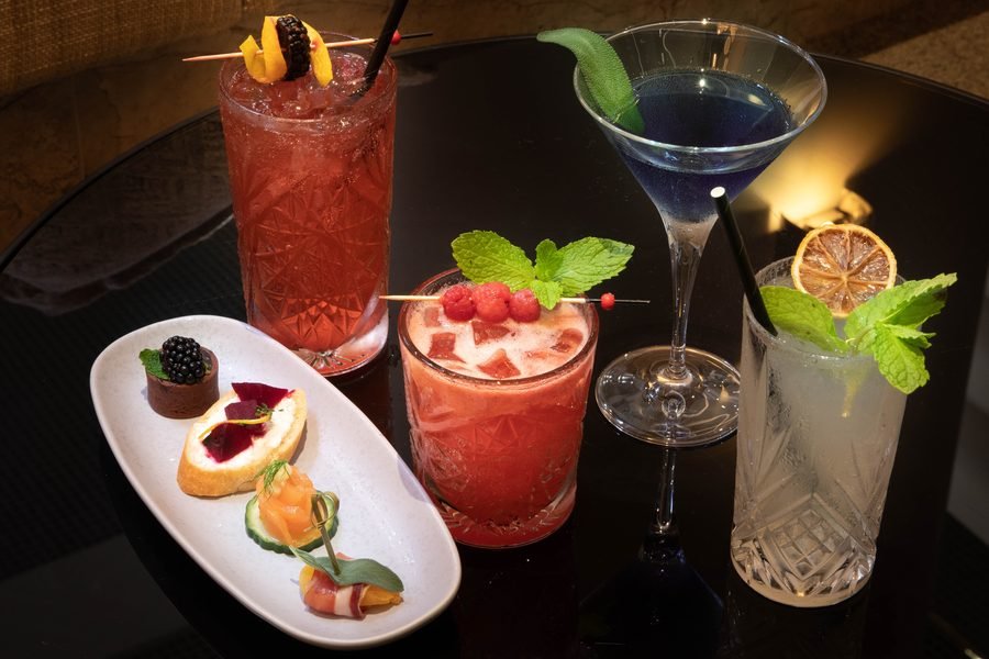 Three cocktails and a plate of canapes styled on a dark bar top